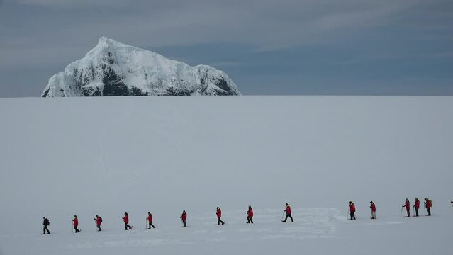 Travel to Antarctica. Tourists in red on snowshoes. Skiers in the background of the Antarctic landscape. People in red suit skiing in the snowy mountains. Journey and Sport Lifestyle.