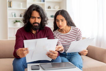 Upset young indian couple checking bills, sitting on couch