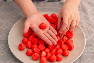 Sweet moments unfold as a child's hands eagerly grasp red fruit candies from a plate, radiating the joy of indulging in chewy treats