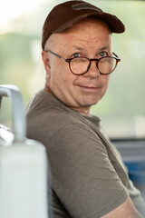 a positive adult man 60+ years old, of European appearance in glasses and a cap, in casual clothes sitting in public transport, looking into the camera, close-up