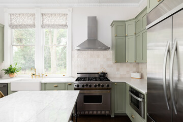 A kitchen detail with sage green cabinets, a gold faucet on an apron sink, brown tiled backsplash,...