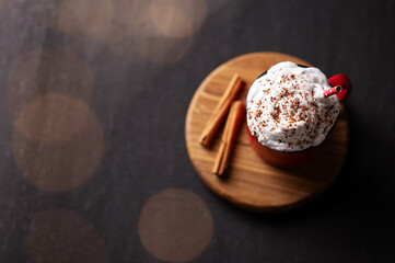 Red mug with hot chocolate or cocoa with whipped cream on a wooden stand with cinnamon sticks  on a dark background with a garland in blur.