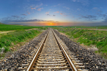 Fototapeta premium Railway stretching through greenery. Setting sun, sunset with blue cloudy sky landscape in the background