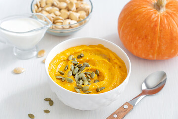 Creamy traditional healthy pumpkin soup puree made with ripe squash mashed with dairy cream and broth decorated with seeds served hot in bowl with spoon and ingredients on white wooden background