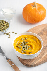 Creamy traditional organic pumpkin soup made of ripe squash mashed or pureed with dairy cream and broth decorated with seeds served in bowl on chopping board with spoon on white wooden table