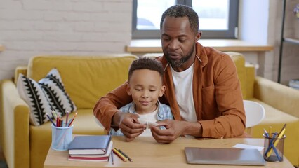 Loving african american father teaching his little son to make origami, making paper butterfly, boy enjoying it at home