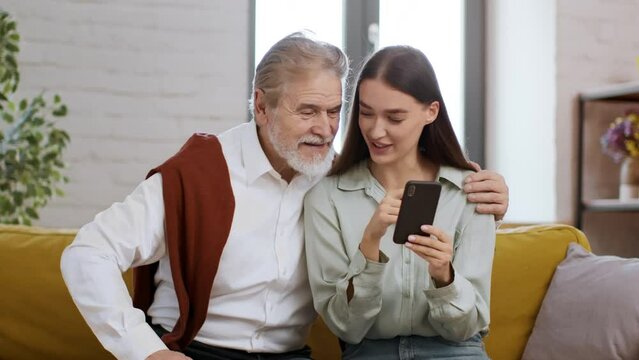 Carefree young lady spending time with her grandfather, reading news on smartphone and discussing them