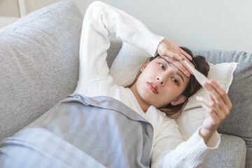 Sick, influenza asian young woman, girl have a fever, hand holding thermometer, touching forehead for check measure body temperature, illness while lying rest on sofa at home. Health care concept.