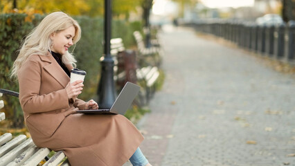 Fototapeta premium Freelance work. Happy woman. Virtual connection. Smiling lady sitting bench in park alley typing laptop holding cup of coffee copy space.