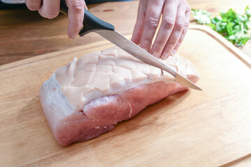 Rind from a raw roast pork is incised in a diamond shape with a sharp knife on a wooden kitchen...