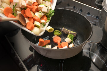 Diced vegetables are poured into an oval casserole pot on the stove top to roast them for a sauce,...
