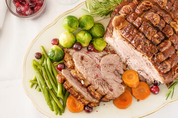 Roast pork with crust as Christmas ham with vegetables and cranberries on a white serving plate, holiday dinner, high angle view from above, copy space, selected focus