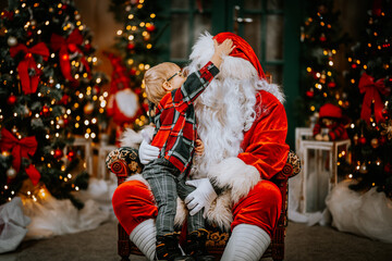 Santa Claus with a sweet child on his lap listens to Christmas wishes while sitting in a chair.