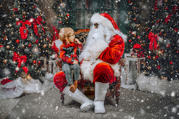 Santa Claus with a sweet child on his lap listens to Christmas wishes while sitting in a chair.