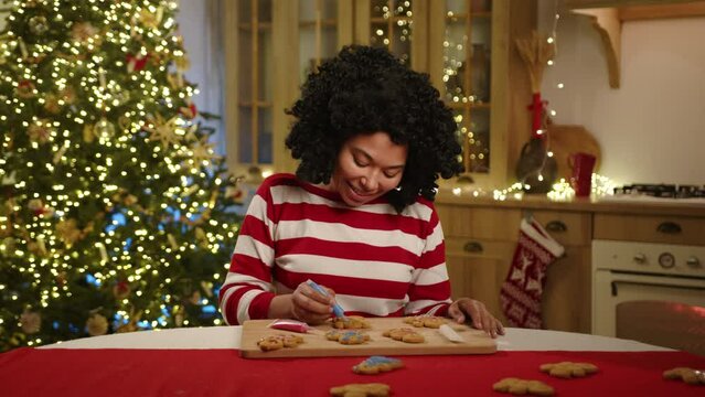 Laughing black woman looking at camera after painting gingerbread man in blue. Modern light kitchen with Christmas stockings, xmas tree with garlands. High quality 4k footage