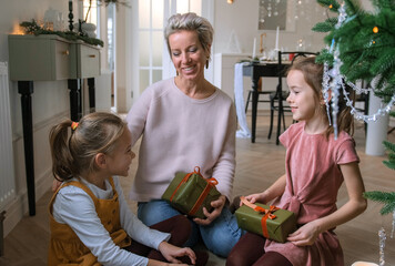 Cheerful mom and her cute daughters exchange Christmas gifts sitting on the floor near the Christmas tree