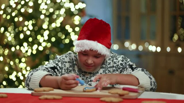 Kid in sweater with winter ornament sitting in festively decorated kitchen, painting gingerbread man, tasting food coloring, looking at camera like angel. High quality 4k footage