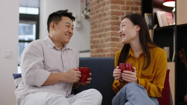 Loving asian man and woman talking with coffee cups at home, discussing news and laughing, tracking shot, slow motion