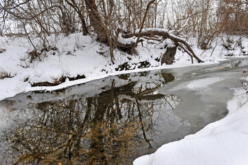 View of a partly frozen river with visible flowing, dark water and banks covered with white snow in...