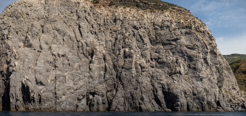 Weathered seaside rock face texture with parts of green and blue water. Aged volcanic stone wall surface background pattern with cracks and scratches. Banner. Ischia Island, Italy.