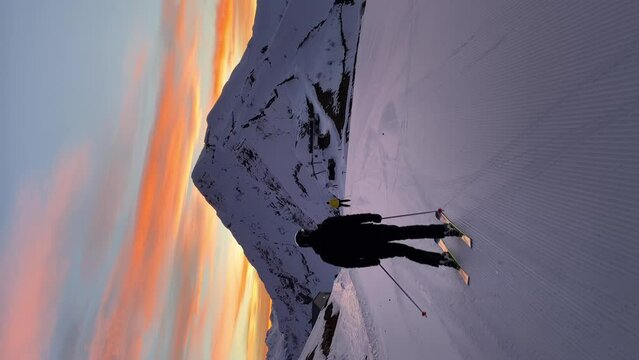 Two skiers smoothly accelerate along a perfectly prepared track at dawn. There is no one at the ski resort yet. Sun has not yet come out from behind the mountain. Golden light reflects off the clouds