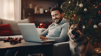 A middle-aged man works on a laptop while sitting in a cozy Christmas living room with a dog on the...