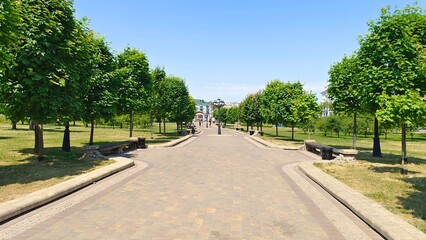 In the city park, paving stone walkways with curbs are laid among mowed lawns and various trees. There are metal poles with lanterns on them. Behind the park are buildings. Sunny summer weather