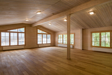 A room with an open space in a luxurious country house made of wooden beams. Unfurnished. Windows with a view of the summer landscape.