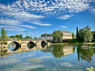 view of a stone bridge in a small French town over a river on a summer day with some white clouds
