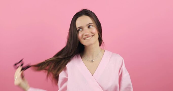 Lovely coquettish brunette woman, winking playfully, flirting. Woman fix her hair isolated on pink color. Brunette hair fashion model face smiling removes hair clipper and untangles hair.