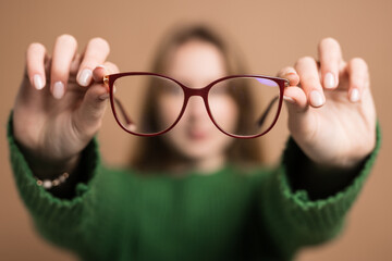 Poor vision concept, Young girl with poor vision holds glasses with hands forward on beige background - obrazy, fototapety, plakaty