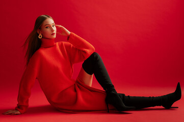 Fashionable confident woman wearing trendy red long knitted turtleneck dress, over the knee high heel boots, posing on red background. Full-length studio fashion portrait. Copy, empty space for text - obrazy, fototapety, plakaty
