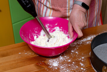 Chef with mixer in hands cooking sweet cheesecake with cooking ingredients on kitchen table
