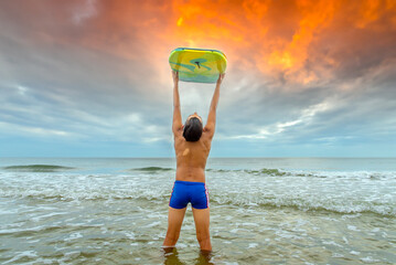 Male child swimmer at the beach holding his boogie board above his head during dramatic sunset