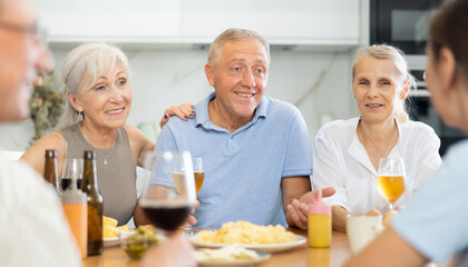 Group of joyful seniors at family dinner drinking and chatting at table in kitchen