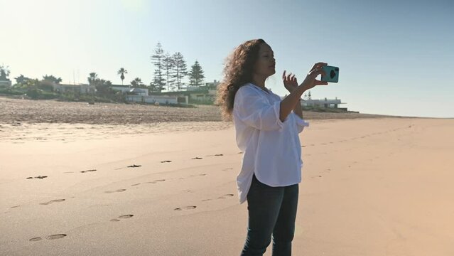 Woman in casual clothing, standing on the sandy beach by the Atlantic ocean, taking photos of the cold and stormy waves crashing against the shore with her smartphone. Travel, tourism, and adventure.
