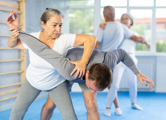 Fototapeta premium Elderly woman twists the arm of attacking man with painful hold in gym. Self-defense lesson