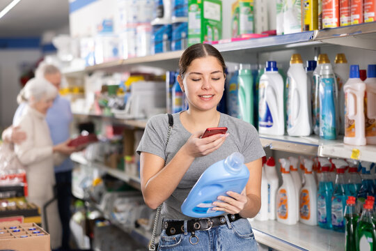 Watchful young woman purchaser taking photo of label of liquid detergent during shopping in a supermarket