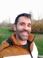 selfie of black haired caucasian man smiling with short beard and orange and white jacket in a park - 684373723