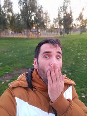 Selfie of Caucasian man with short black hair and brown and white coat covering his mouth with his hand surprised in park during autumn or winter - 684373735