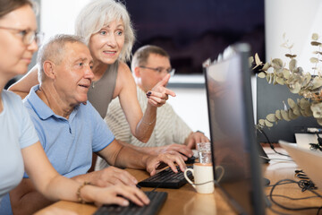 Elderly people help each other learn how to use computer. Woman suggests right decision to an mature man - obrazy, fototapety, plakaty