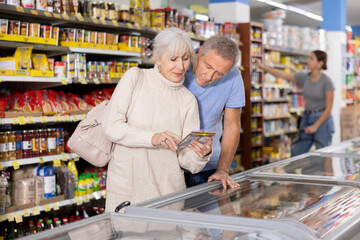 Watchful aged man and woman customers selecting frozen food from market fridge in superstore