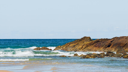Australian coast with volcanic rocks at the shore, view from the beach to the horizon with blue water with waves on a summer sunny day.