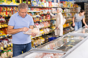 Positive mature man shopping in supermarket, choosing frozen convenience food
