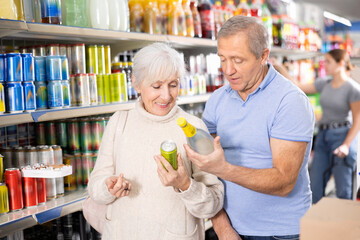 Positive elderly couple standing in aisle of supermarket, friendly discussing while choosing...