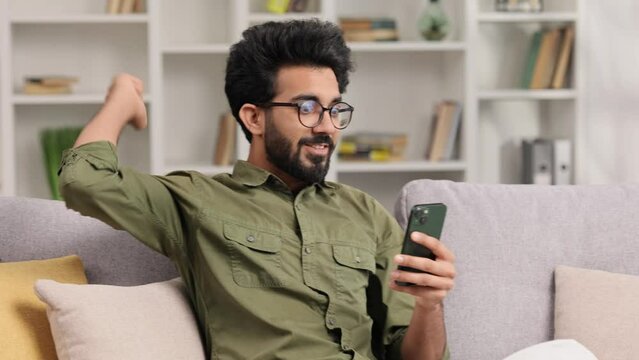 Satisfied Indian man in black glasses checking his email and happy with good news. A young man winner celebrating the victory sitting on the couch, raises his hands uphill.