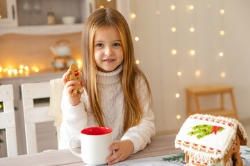 Cute little girl in white wool sweater holding decorated Christmas gingerbread men cookie in a...