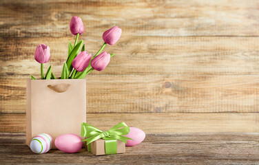  paper bag contains a bouquet of pink tulips and multicolored eggs with a gift box on a wooden background with a place for text