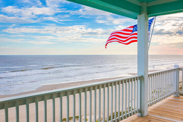 View of the ocean and sandy beach under a blue sky with light white clouds from a wooden deck balcony with an American flag
