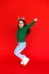 Happy little girl in reindeer horns with Christmas gift on red background. A little girl has fun on a red isolated background. A child in a warm green sweater holds a gift box.